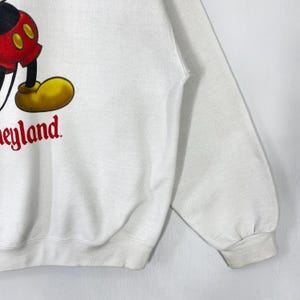 Puede incluir: Sudadera blanca con una imagen parcial de Mickey Mouse, con pantalones cortos rojos, piernas negras y zapatos amarillos. La palabra "neyland" est&aacute; impresa en rojo. La sudadera tiene cuello redondo y mangas largas.