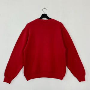 May include: A plain red sweatshirt with long sleeves, suspended from a black hanger against a white backdrop. The rear of the garment is displayed, showcasing a simple, classic design. The material seems to be of medium weight, appropriate for cooler temperatures.