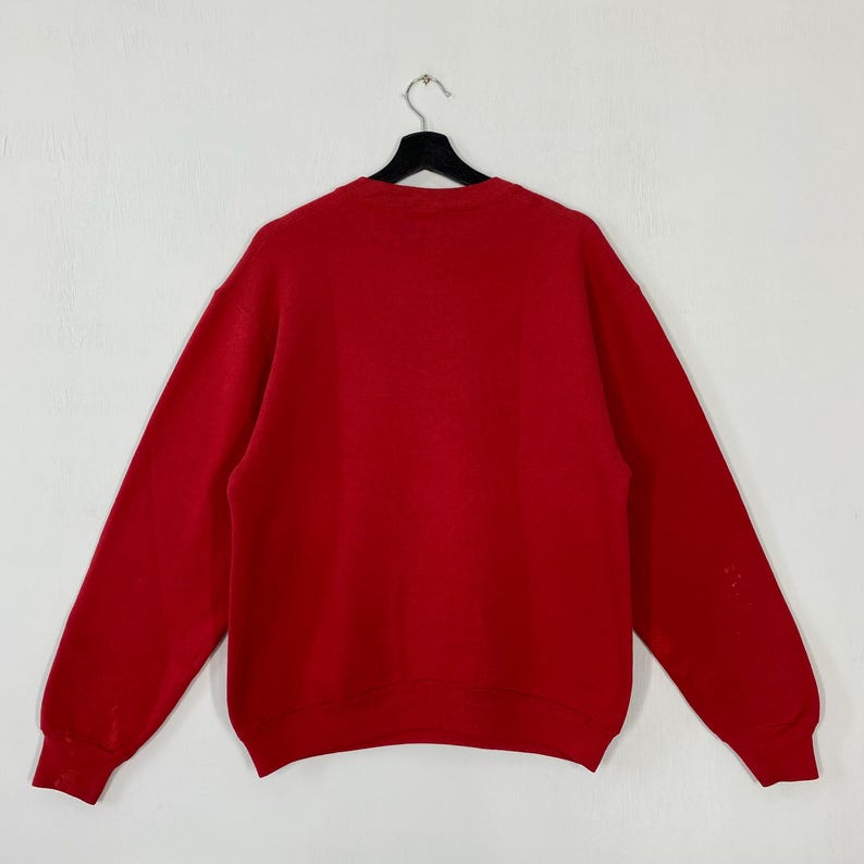 May include: A plain red sweatshirt with long sleeves, suspended from a black hanger against a white backdrop. The rear of the garment is displayed, showcasing a simple, classic design. The material seems to be of medium weight, appropriate for cooler temperatures.