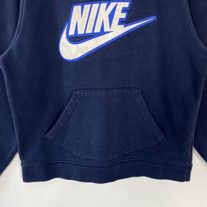 May include: Navy blue hoodie with a kangaroo pocket. The Nike logo is prominently displayed in white with a blue outline on the upper chest. The sweatshirt has a drawstring hood and ribbed cuffs and hem.