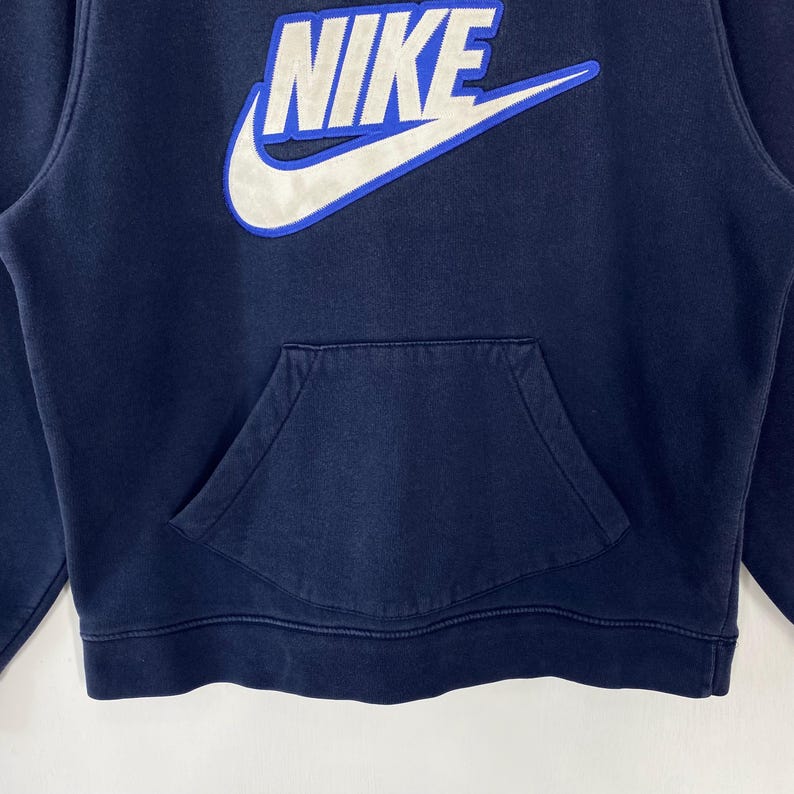 May include: Navy blue hoodie with a kangaroo pocket. The Nike logo is prominently displayed in white with a blue outline on the upper chest. The sweatshirt has a drawstring hood and ribbed cuffs and hem.