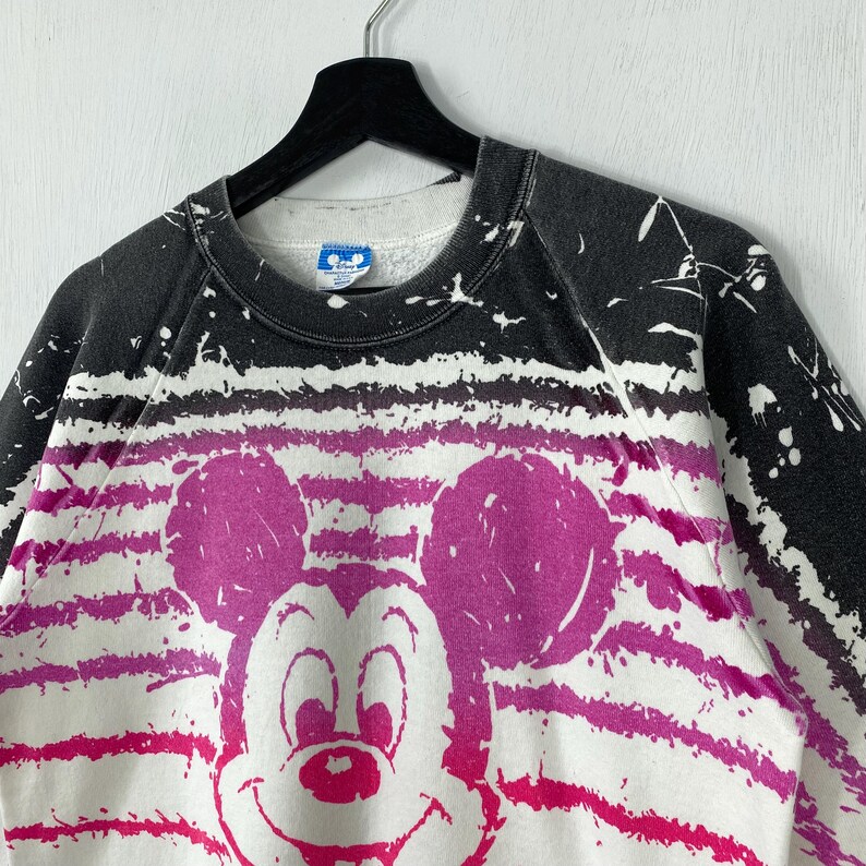 May include: A vintage crew neck sweatshirt featuring a large Mickey Mouse graphic in shades of pink and white. The top portion is black with white paint splatters. The design includes horizontal stripes and is hanging on a black hanger.