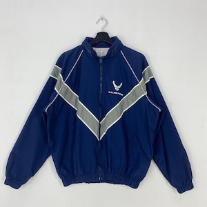 May include: A navy blue zip-up windbreaker jacket featuring a white U.S. Air Force logo on the chest. The jacket has a high collar, long sleeves with elastic cuffs, and a prominent grey chevron stripe pattern across the front and shoulders.