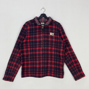 May include: Red and black plaid fleece pullover with a half-zip closure. The long-sleeved shirt features a small logo patch on the chest and a gray collar. The North Face brand is visible on the logo patch.