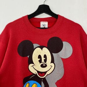 May include: Red crew neck sweatshirt with a large Mickey Mouse graphic. The design features Mickey's face and ears in black, with a faded grey outline. A small Mickey Mouse logo tag is near the neckline.
