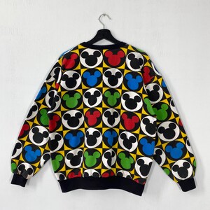 May include: A black, yellow, and white sweatshirt with a repeating pattern of Mickey Mouse heads in red, green, blue, and black. The sweatshirt has a black collar and cuffs. The background is a checkerboard pattern.