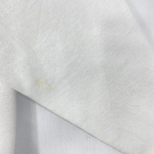 Puede incluir: Primer plano de una tela blanca con una mancha marr&oacute;n claro. La tela parece ser un material suave, posiblemente una sudadera o una prenda similar. La mancha est&aacute; ubicada en el lado inferior izquierdo de la imagen.