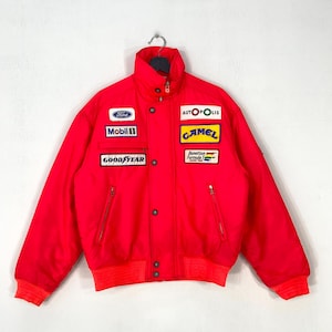 May include: A bright red racing jacket featuring a high collar, long sleeves, and ribbed cuffs. The front displays various embroidered patches including Ford, Mobil 1, Goodyear, Autopolis, Camel, and Benetton Formula 1 Racing Team logos.