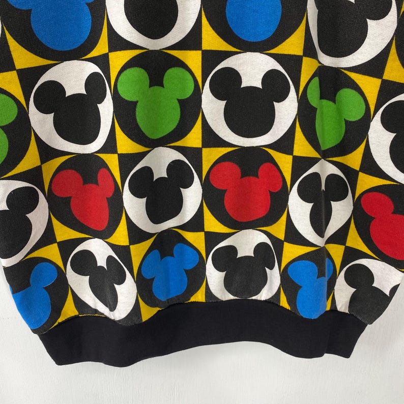 May include: Black sweatshirt with a repeating pattern of Mickey Mouse heads in various colors, including black, white, red, green, and blue. The design is set against a yellow and black checkered background. The bottom hem and cuffs are solid black.