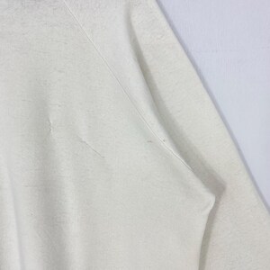 Pu&ograve; includere: Primo piano di una felpa color crema. Il tessuto mostra segni di usura con piccoli strappi e imperfezioni. Le cuciture delle maniche e delle spalle sono visibili, indicando la costruzione del capo. L'aspetto generale suggerisce uno stile casual e usurato.