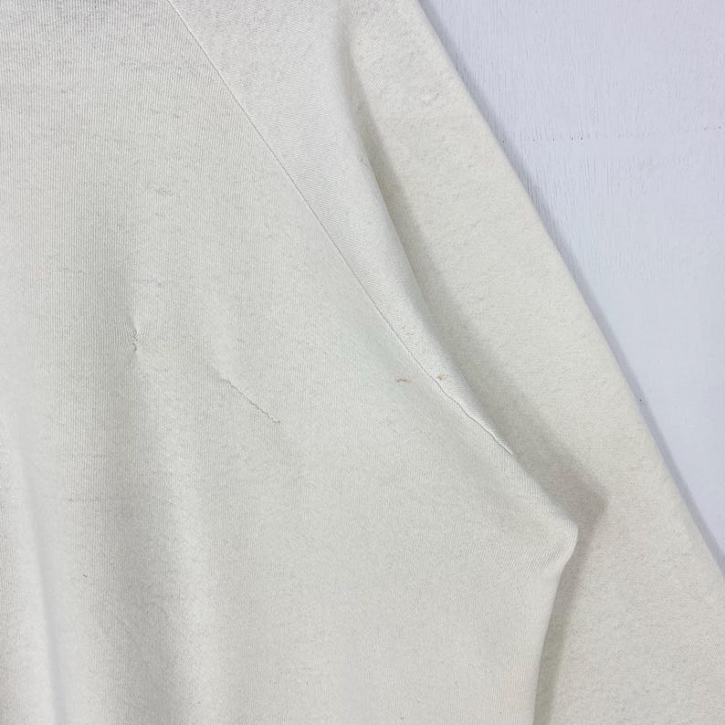 Pu&ograve; includere: Primo piano di una felpa color crema. Il tessuto mostra segni di usura con piccoli strappi e imperfezioni. Le cuciture delle maniche e delle spalle sono visibili, indicando la costruzione del capo. L'aspetto generale suggerisce uno stile casual e usurato.