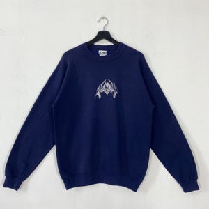 Puede incluir: Sudadera azul marino con un dise&ntilde;o gr&aacute;fico blanco en la parte delantera. La parte superior de manga larga con cuello redondo est&aacute; colgada de una percha negra sobre un fondo blanco. El dise&ntilde;o es un emblema estilizado.