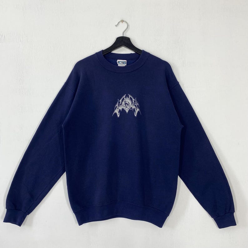 Puede incluir: Sudadera azul marino con un dise&ntilde;o gr&aacute;fico blanco en la parte delantera. La parte superior de manga larga con cuello redondo est&aacute; colgada de una percha negra sobre un fondo blanco. El dise&ntilde;o es un emblema estilizado.