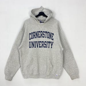 May include: A light gray hoodie with the text "CORNERSTONE UNIVERSITY" in navy blue across the chest. The hoodie has a front pocket, a drawstring hood, and a small logo on the lower right. The sweatshirt is hanging on a black hanger.