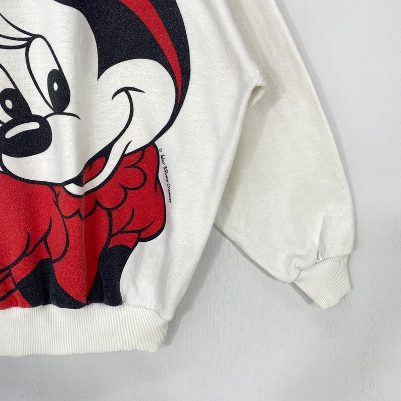 Puede incluir: Sudadera blanca con un gran gr&aacute;fico de Mickey Mouse. El dise&ntilde;o incluye a Mickey sonriendo con detalles en rojo y negro. La manga y el pu&ntilde;o son visibles. El copyright de The Walt Disney Company est&aacute; impreso en el lateral.