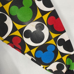 May include: Close-up of a fabric with a repeating pattern of Mickey Mouse heads in black, red, green, and blue, set against a black background with yellow diamond shapes. The Mickey Mouse heads are inside white circles.