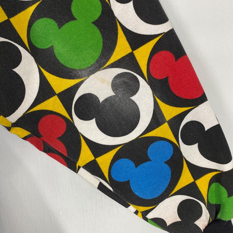 May include: Close-up of a fabric with a repeating pattern of Mickey Mouse heads in black, red, green, and blue, set against a black background with yellow diamond shapes. The Mickey Mouse heads are inside white circles.