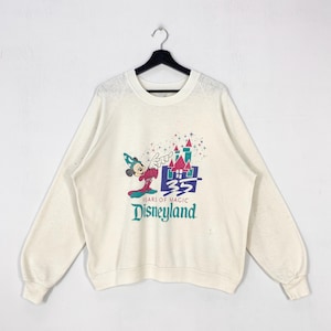 Pu&ograve; includere: Felpa color crema con una grafica colorata di Topolino con cappello da mago, un castello e la scritta "35 Years of Magic Disneyland". La maglia a maniche lunghe &egrave; appesa a una gruccia nera.