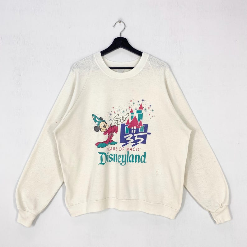 Pu&ograve; includere: Felpa color crema con una grafica colorata di Topolino con cappello da mago, un castello e la scritta "35 Years of Magic Disneyland". La maglia a maniche lunghe &egrave; appesa a una gruccia nera.