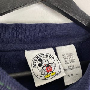 May include: Navy blue sweatshirt with a Mickey & Co. label featuring Mickey Mouse in red, yellow, and white. The label also includes the Disney logo and fabric content details. The size is labeled as L.