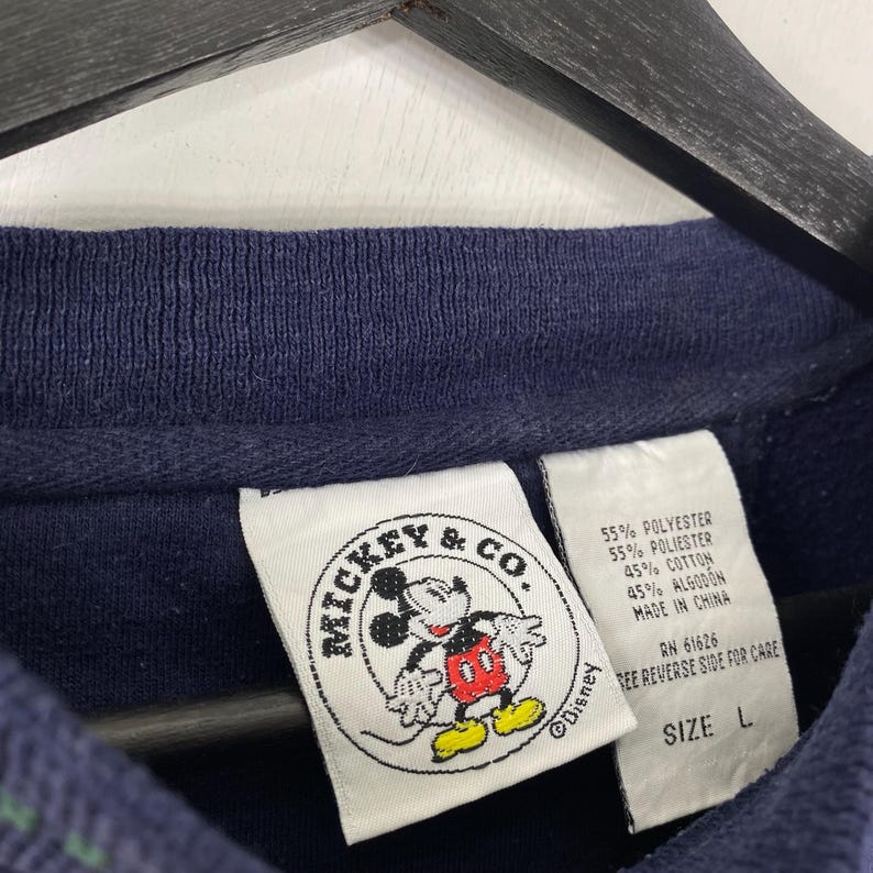 May include: Navy blue sweatshirt with a Mickey & Co. label featuring Mickey Mouse in red, yellow, and white. The label also includes the Disney logo and fabric content details. The size is labeled as L.