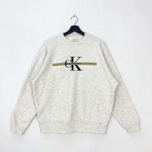 Peut inclure: Un sweat-shirt Calvin Klein Jeans gris clair. Le sweat-shirt présente le logo Calvin Klein en noir avec une ligne horizontale dorée. Il est suspendu à un cintre noir sur fond blanc.