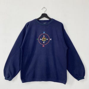 May include: Navy blue sweatshirt with a crew neck and long sleeves. The front features an embroidered compass design with the letter "M" at each point. The compass design is surrounded by colorful floral and butterfly accents.