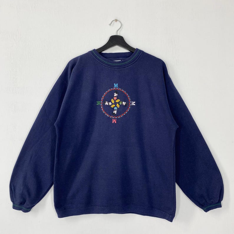 May include: Navy blue sweatshirt with a crew neck and long sleeves. The front features an embroidered compass design with the letter "M" at each point. The compass design is surrounded by colorful floral and butterfly accents.