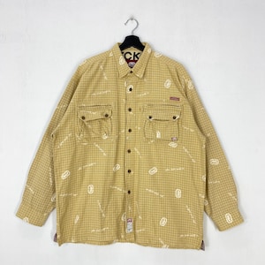 May include: A long-sleeve button-down shirt in a mustard yellow color with a white grid pattern and scattered white logo prints. The shirt features two chest pockets with zipper closures, a standard collar, and a button-up front placket.