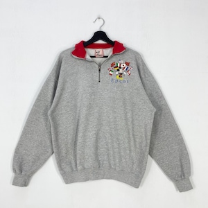 May include: A gray sweatshirt with a red collar and a quarter-zip closure. The sweatshirt features an embroidered design of Mickey Mouse, flags, and the word "Epcot". The long-sleeved top is hanging on a black hanger.