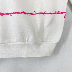 May include: White sweatshirt with a horizontal pink splatter design across the chest. The bottom hem and cuffs are ribbed. The fabric appears to be a soft, comfortable material.