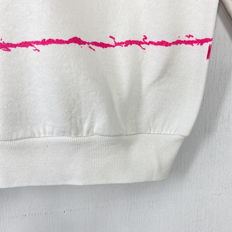 May include: White sweatshirt with a horizontal pink splatter design across the chest. The bottom hem and cuffs are ribbed. The fabric appears to be a soft, comfortable material.