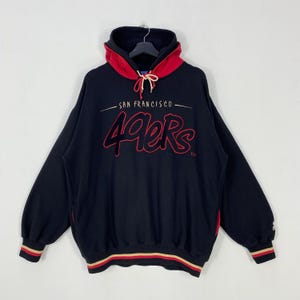 May include: Black hoodie with a red-lined hood and cuffs. The front features the text "SAN FRANCISCO" above the red "49ers" logo. The cuffs have red, gold, and black stripes. The hoodie is hanging on a black hanger.