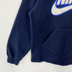 May include: Navy blue hoodie with a white and blue Nike logo on the upper chest. The hoodie features a kangaroo pocket and ribbed cuffs and hem. The sleeve is visible on the left side of the image.