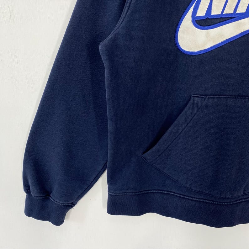 May include: Navy blue hoodie with a white and blue Nike logo on the upper chest. The hoodie features a kangaroo pocket and ribbed cuffs and hem. The sleeve is visible on the left side of the image.