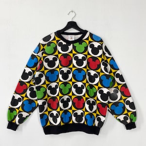 Puede incluir: Una sudadera negra, amarilla y blanca con un patrón repetido de cabezas de Mickey Mouse en rojo, verde y azul. La sudadera tiene cuello y puños negros, y está colgada de una percha negra.