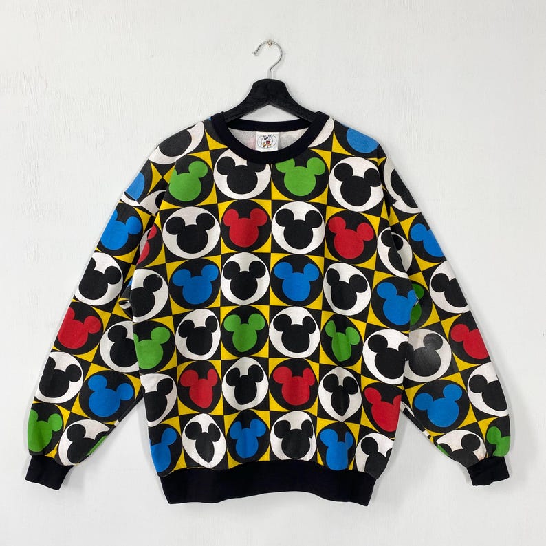 May include: A black crew neck sweatshirt with a colorful Mickey Mouse pattern. The design features Mickey Mouse heads in white, red, green, and blue, set against a yellow and black checkered background. The cuffs and collar are black.