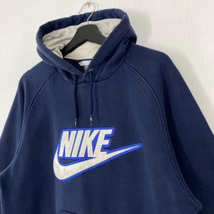 May include: Navy blue Nike hoodie with a white and blue logo. The hoodie has a drawstring hood and a kangaroo pocket. The Nike logo is embroidered on the front. The interior of the hood is light gray.