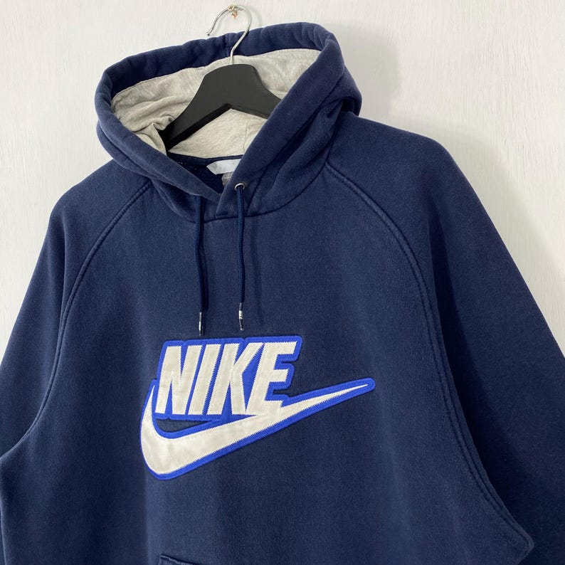 May include: Navy blue Nike hoodie with a white and blue logo. The hoodie has a drawstring hood and a kangaroo pocket. The Nike logo is embroidered on the front. The interior of the hood is light gray.