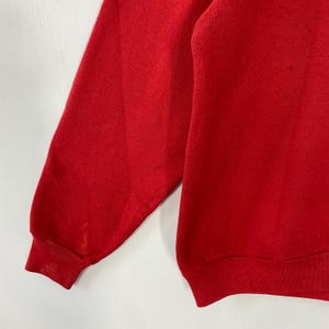 May include: A close-up view of a red sweatshirt. The image focuses on the sleeve and part of the body of the garment. The fabric looks soft, possibly fleece-lined. There are some minor stains on the sleeve.