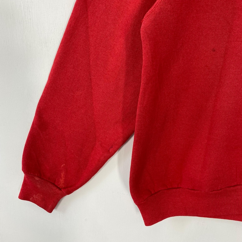 May include: A close-up view of a red sweatshirt. The image focuses on the sleeve and part of the body of the garment. The fabric looks soft, possibly fleece-lined. There are some minor stains on the sleeve.