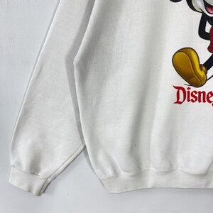 Puede incluir: Sudadera blanca con un gran gr&aacute;fico de Mickey Mouse y la palabra "Disney" en rojo. La sudadera tiene mangas largas y cuello redondo. Un dise&ntilde;o cl&aacute;sico de Disney.