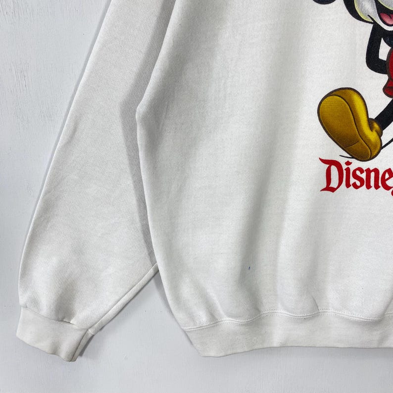 Puede incluir: Sudadera blanca con un gran gr&aacute;fico de Mickey Mouse y la palabra "Disney" en rojo. La sudadera tiene mangas largas y cuello redondo. Un dise&ntilde;o cl&aacute;sico de Disney.
