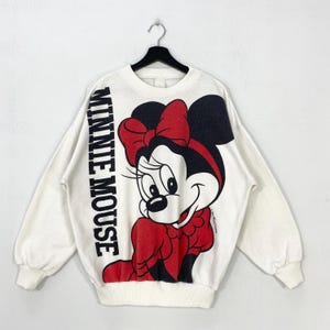 Puede incluir: Sudadera blanca con un gran gr&aacute;fico de Minnie Mouse con un lazo y un vestido rojos. Las palabras "MINNIE MOUSE" est&aacute;n impresas verticalmente en la manga izquierda. La sudadera est&aacute; colgada de una percha negra.