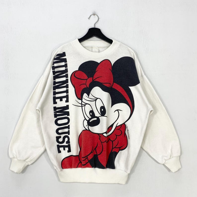 Puede incluir: Sudadera blanca con un gran gr&aacute;fico de Minnie Mouse con un lazo y un vestido rojos. Las palabras "MINNIE MOUSE" est&aacute;n impresas verticalmente en la manga izquierda. La sudadera est&aacute; colgada de una percha negra.