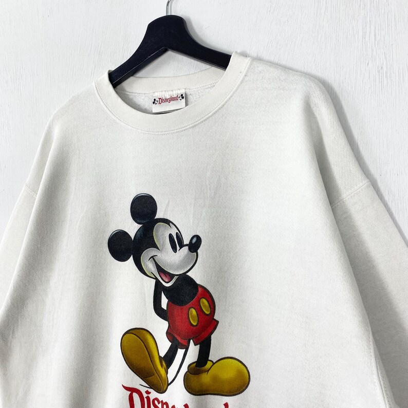 Puede incluir: Sudadera de color crema con un gran gr&aacute;fico de Mickey Mouse en negro, rojo y amarillo. La sudadera tiene cuello redondo y una peque&ntilde;a etiqueta de Disneyland. El logotipo de Disneyland est&aacute; impreso en rojo en la parte inferior.