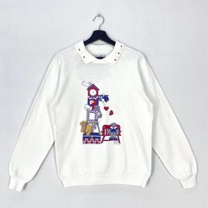Könnte beinhalten: Ein weißes Langarm-Sweatshirt mit einem Kragen, der mit kleinen roten Herzakzenten verziert ist. Die mittlere Grafik zeigt einen Stapel Spielzeug, darunter einen Uhrturm, eine Gans, einen Teddybären und eine rote Bank mit einem Bären.