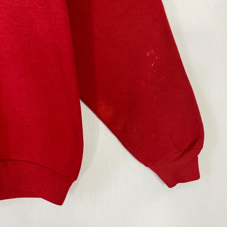 May include: A close-up of a red sweatshirt sleeve. The sleeve has a ribbed cuff and some discolouration on the upper arm. The background is a plain white surface.