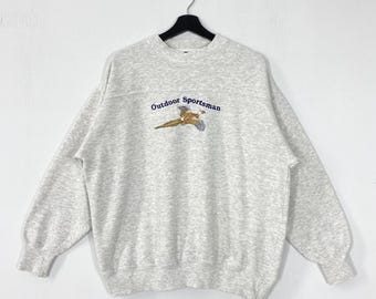 May include: Light gray sweatshirt with the text "Outdoor Sportsman" above an embroidered pheasant in flight. The long-sleeved crew neck top is made of a soft material and is suitable for casual wear.