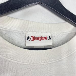 Puede incluir: Primer plano de un cuello de sudadera blanco con una etiqueta de Disneyland. La etiqueta presenta el logotipo de Disneyland en rojo y negro, con siluetas de Mickey Mouse. La sudadera tiene un cuello acanalado.